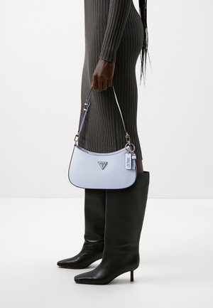 Ljusblå handväska med texturerad yta, svart kantning och metalldetaljer i form av logotyp. Väskan har axelrem och hålls av en person klädd i grå ribbstickad klänning och svarta stövlar.