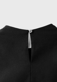 Gros plan sur l'encolure d'un vêtement noir avec un petit bouton gris sécurisant une ouverture en forme de trou de serrure à l'arrière.