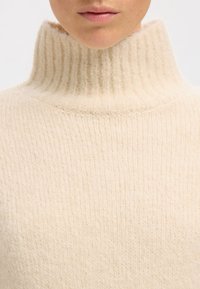 Kremo spalvos golfas megztas iš minkštos, taip pat riboto audinio, pasižymintis aukštu apykakle ir struktūruota apdaila.