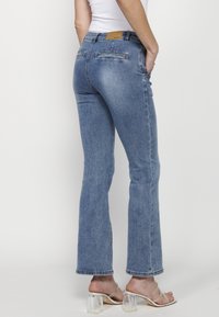 Blå jeans med utsvängda ben och medelhög midja, utrustade med två bakfickor, diskret blekning och en ren fåll. Bärs med klara högklackade sandaler.