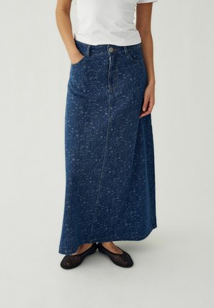 Moteris dėvi ilgą mėlyną džinsinę sijoną su subtiliu nusidėvėjimo raštu, baltą trumpomis rankovėmis marškinėlį ir juodus tinklinio rašto butučius batus, stovinti su rankomis kišenėse.