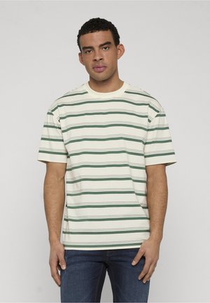 Mann trägt cremefarbenes T-Shirt mit kurzen Ärmeln und grünen horizontalen Streifen sowie dunkelblaue Jeans, steht vor einem einfarbigen grauen Hintergrund.