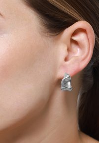 Silberne Metallohrringe in abstrakter Form mit glatter Textur, die am Ohrläppchen befestigt sind. Die reflektierende Oberfläche verstärkt das Design.