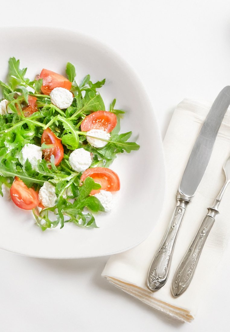 Insalata di rucola con pomodori a metà e ciuffi di formaggio in una ciotola bianca, accompagnata da posate in argento su un tovagliolo di crema piegato.