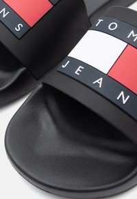 Black rubber slides with a wide strap featuring the text "TOMMY JEANS" and color block design in red, white, and blue. Smooth texture.