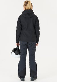 Zwarte skijack met capuchon, gecombineerd met zwarte sneeuwbroek. De persoon houdt een witte skihelmet vast. Kenmerken zijn het slanke ontwerp en weerbestendige materialen.