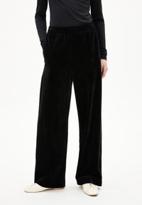 Person, die schwarze, weit geschnittene Samenhosen, ein schwarzes Langarmoberteil und cremefarbene flache Schuhe trägt, steht mit den Händen in den Taschen.