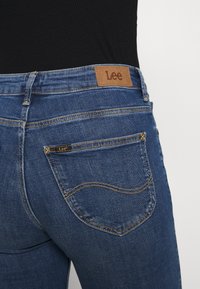 Jeans de mezclilla azul con un parche de cuero en la parte trasera etiquetado "Lee". Presenta costuras amarillas en contraste y dos bolsillos traseros con costuras curvas.