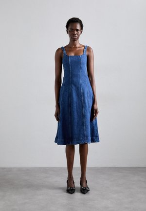 Denim A-line mekko, jossa on neliön muotoinen kaula-aukko, ohuet hihnat ja näkyvät saumat. Polvien ylle ulottuva, avara helma tummansinisessä. Mustat korkokengät.