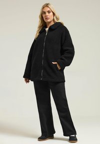 Musta fleece-takki, jossa on kauluspanta, etuvetoketju ja sivutaskut; päällä mustat housut ja mustat kengät, joissa on teksturoitu pohja.