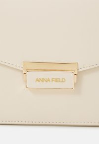 Clutch in ecopelle color crema con chiusura in metallo dorato incisa con il nome "ANNA FIELD." Texture liscia e linee pulite.