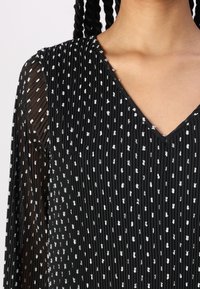 Svart plisserad blus med V-ringning, prydd med vita sporadiska prickar. Lätt väv med genomskinliga ärmdetaljer och texturerat material.