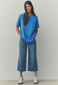 Camiseta de algodón azul con mangas cortas combinada con culottes de denim desgastado de pierna ancha y zapatillas negras y blancas, de pie sobre una superficie clara.