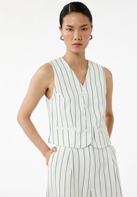 Junge Frau mit zurückgegeltem Haar, die eine weiße ärmellose Weste mit Streifen und passende hoch taillierte Streifenhose trägt, mit Händen in den Taschen.