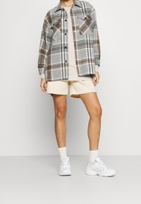 Szara koszulowa kurtka w kratę z brązowymi i białymi akcentami, beżowy t-shirt, beżowe szorty oraz białe trampki z fakturowaną podeszwą i jasnymi skarpetkami.