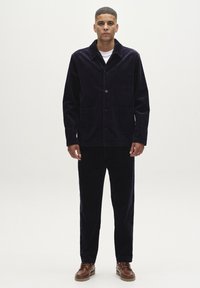 Navy corduroy jas met een puntige kraag, knoopsluiting aan de voorkant en twee voorzakken, gecombineerd met bijpassende corduroy broek en bruine schoenen.