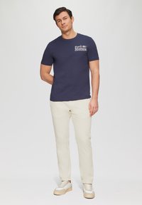 Navyblauwe katoenen t-shirt met het "Ford Mustang" logo in het wit, gecombineerd met crème kleurige broek en lichte sneakers. Casual pasvorm, korte mouwen.