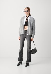 Cardigan grigio a maglia sopra una camicia bianca e un top corto nero, abbinato a jeans svasati scuri. Gli accessori includono una borsa nera e occhiali da sole rettangolari.
