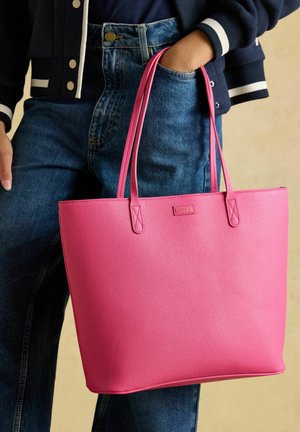 Inimene, kes kannab tumedat jopet ja siniseid teksapükse, hoides suurt roosat tekstuuriga käekotti ühe käega sangadest, samal ajal kui teine käsi on taskus.