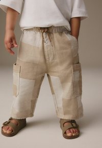 Beige Patchwork-Hosen mit elastischem Bund und Kordelzug, ausgestattet mit Seitentaschen. Kombiniert mit braunen Sandalen und einem weißen kurzärmligen Shirt.