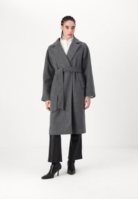 Grau-wollner Mantel mit einem taillierten Gürtel, Reverskragen und langen Ärmeln, kombiniert mit schwarzen Lederhosen und spitzen Knöchelstiefeln.