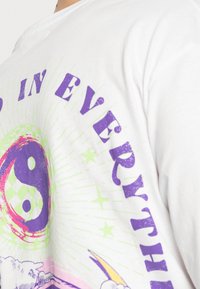 Camiseta blanca de algodón con un diseño gráfico colorido que incluye un símbolo de yin-yang, estrellas y el texto "EN TODO" en púrpura y verde.