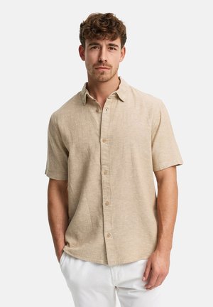 Młody mężczyzna z kręconymi włosami stoi zwrócony przodem, ubrany w beżową koszulę na krótki rękaw z guzikami i białe spodnie, z rękami w kieszeniach.