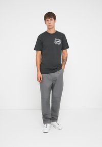 Black cotton t-shirt with a white graphic pocket design, paired with gray pants and white sneakers, standing against a plain background.