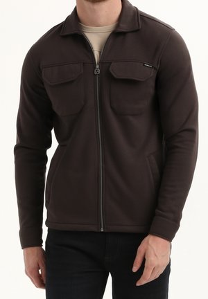 Mann trägt eine dunkelbraune Jacke mit Reißverschluss, die Brusttaschen mit Klappen hat, und darunter ein beiges Hemd, steht vor einem schlichten Hintergrund.