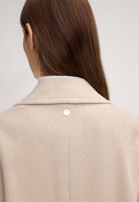 Beige wollen jas met een geknipte kraag, voorzien van een zilveren ronde sluiting aan de achterkant. De stof lijkt zacht met een gladde textuur.