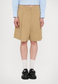 Khakifarbene High-Waisted-Shorts mit Falten, aus strukturiertem Stoff. Kombiniert mit schwarzen Schuhen und weißen Socken, getragen mit einem hellblauen Oberteil.