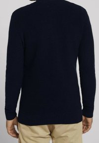 Pull en maille bleu marine foncé avec texture côtelée, manches longues et ourlet droit. La vue de dos met en valeur un détail de couture horizontal.