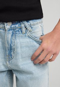 Hand resting in front pocket of light blue denim jeans worn with a black ribbed top against a plain background.