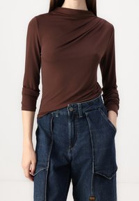 Mörkbrun långärmad topp med en krage i cowl-stil och samlad detalj, i kombination med lösa blå denim cargo-byxor med sidfickor och knappstängning.