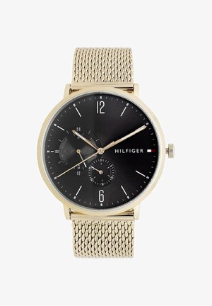 Rund guldfarvet armbåndsur med sort urskive, guldfarvet mesh-rem, tre visere, dato og 24-timers underskiver samt Hilfiger-logo på højre side.