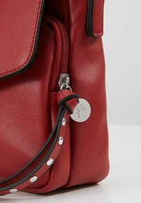 Rudá kožená taška s rohem, stříbrným zipem, černým popruhem se stříbrnými cvočky a kulatým stříbrným logo přívěskem připojeným k zipu.