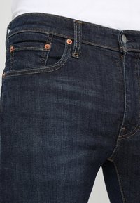 Vaqueros de mezclilla oscura con un corte clásico, que presentan herrajes de cobre, cinco bolsillos y detalles de costuras visibles en color naranja. Textura suave.