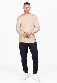 Camicia beige a maniche lunghe realizzata in tessuto morbido, abbinata a pantaloni della tuta blu navy. Il modello indossa sneakers bianche, in piedi su uno sfondo semplice.