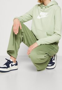 Ljust grön hoodie med vitt Nike-logga, i kombination med matchande olivgröna byxor och marinblå sneakers med vita accent.