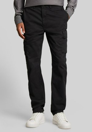 Mann trägt schwarze Cargo-Hosen und weiße Sneakers, steht vor neutralem Hintergrund, Hände in den Taschen.
