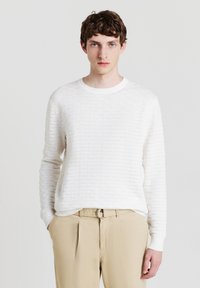 Maglione bianco con texture, collo tondo e maniche lunghe, caratterizzato da un sottile motivo orizzontale. Abbinato a pantaloni beige.