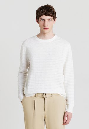 Weißer, strukturierter Pullover mit rundem Kragen und langen Ärmeln, der ein dezentes horizontales Muster aufweist. Kombiniert mit beigen Hosen.