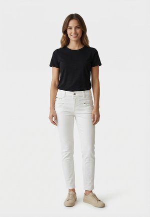 Donna in piedi, rivolta verso l'obiettivo, sorridente, che indossa una maglietta nera a maniche corte, pantaloni bianchi e sneakers beige su uno sfondo chiaro e uniforme.