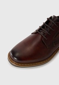 Zapato de vestir de cuero marrón con una textura suave, puntera redondeada, cordones marrones y un detalle de suela cosida. Diseño clásico adecuado para vestimenta formal.