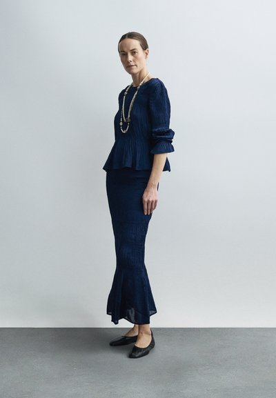 Femeie în picioare, purtând o bluză cu mâneci lungi texturată, de culoare bleumarin, și o fustă lungă asortată, cu pantofi negri și un colier lung de perle.