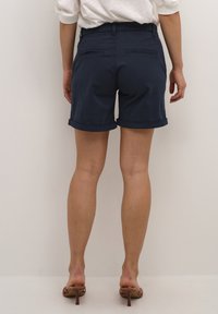 Tmavě modré bavlněné šortky s ohrnutými lemy, se dvěma zadními kapsami a hladkou texturou, doplněné hnědými sandály na podpatku.