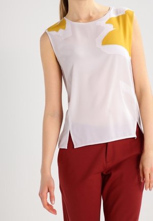 Mouwloze blouse van lichte stof, met een witte basis en abstracte gele en grijze patronen, en zijsplitsen, gecombineerd met bordeauxrode pantalons.