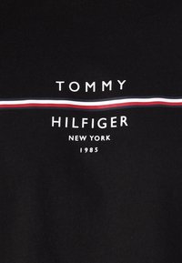 Sort stof med hvid tekst "TOMMY HILFIGER NEW YORK 1985" og en horisontal stribe i hvid, rød og blå på tværs af midten.