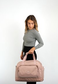 Svetloružová cestovná taška s dvojitými rukoväťami, priestranným dizajnom, prednou zipsovou vreckom a hladkou textúrou látky, držená modelkou v neformálnom oblečení.