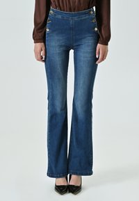 Jean taille haute évasée en denim bleu foncé avec des accents de boutons dorés sur les côtés. Texture lisse et silhouette ajustée au niveau des hanches.